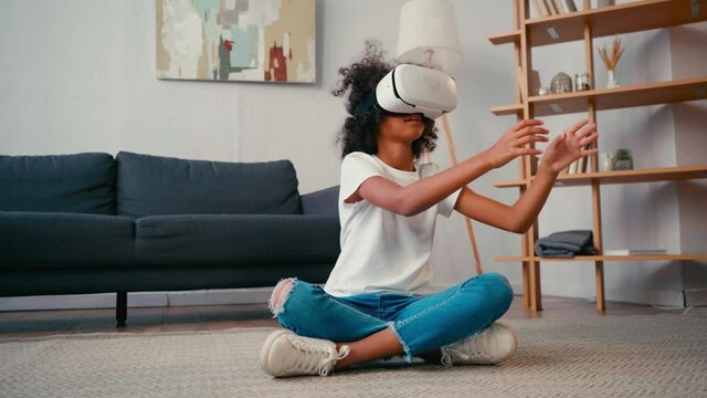 African american girl with crossed legs gesturing while using vr headset at home