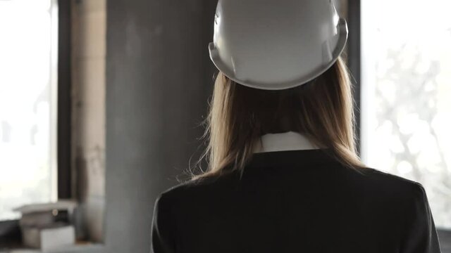 Back view of young woman engineer specialist or worker in a uniform and protective white helmet walking to construction site