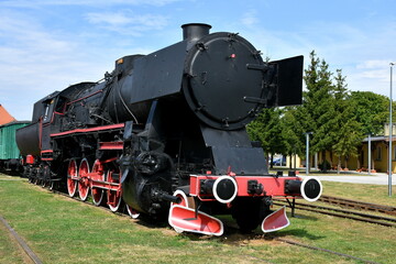 Obraz premium Close up on a vintage train with a black metal body and red and white wheels standing on a side track surrounded with some trees and with a train station visible in the background in Poland