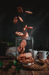 baked cookies falling into a glass jar, on a dark background, with plants and cups of coffee, on rustic wood