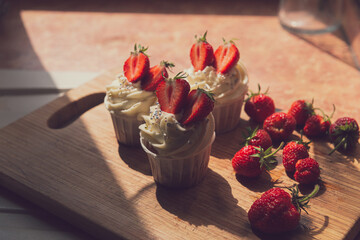 Appetizing cupcakes with white cream decorated with fresh strawberries lit by the rays of the morning sun.