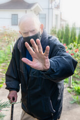 elderly white-haired Caucasian in a black medical mask makes a stop gesture. Being isolated and staying at home is the main requirement of quarantine in the event of a viral pandemic. 