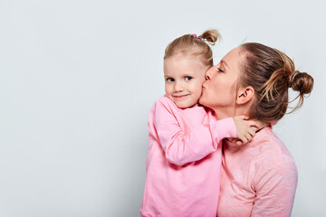 Little daughter hugging and kissing her mother spending time together in pink clothes over gray...