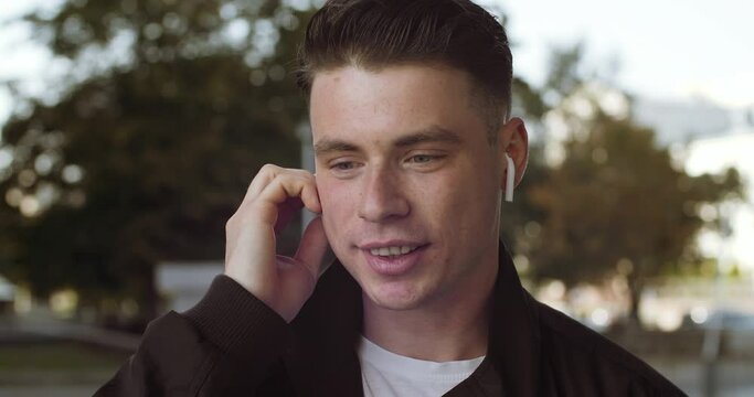 Portrait of young brunette guy man in stylish clothes stands alone outdoors, wears wireless headphones, speaks on phone, listens to interlocutor, answers call, communicates remotely, smiling close-up