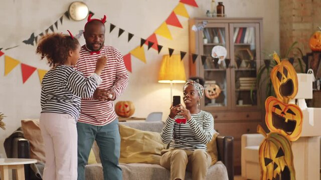 Little black girl throwing ball into boxes decorated with paper jack-o-lanterns and then jumping with arms raised and celebrating victory while playing Halloween game with parents at home