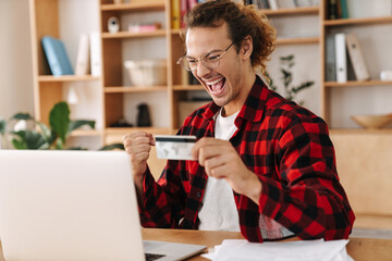 Guy making winner gesture and holding card while working with laptop