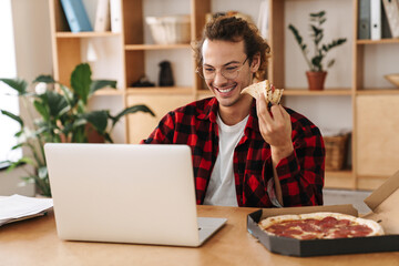 Handsome smiling guy eating pizza and working with laptop at office