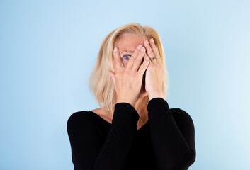 Studio portrait of 60+ woman with blonde page looking through her fingers on light blue background.