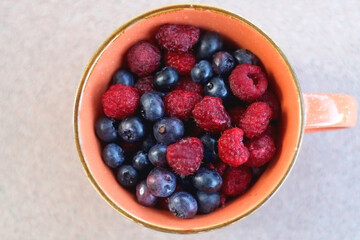 Orange ceramic cup with raspberries and blueberries on a table. Top view.