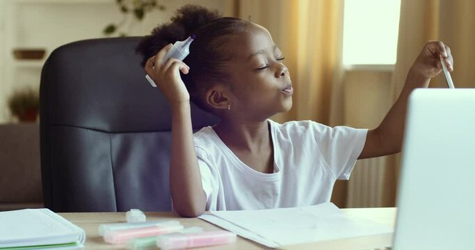 Cute funny little african american girl sits at home at table in front of laptop, draws picture on paper with markers, closes her eyes, chooses random pencil, plays alone, says comments talk on camera