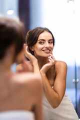 Smiling young woman in bathrobe and towel on head looking at mirror in bathroom