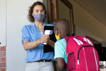 Female teacher wearing a face mask measuring temperature of a boy