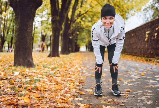 Young fit athletic smiling woman dressed modern running clothes standing and resting on knees on the footway after jogging in the autumnal city park. Active running people concept image.