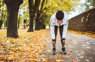 Mid aged fit athletic woman dressed modern running clothes standing and bend over to stretch the back on the footway after jogging in the autumnal city park. Active running people concept image.