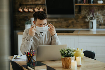 Young freelance worker using face mask while working on laptop at home.