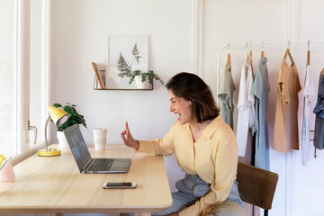 Cheerful young woman showing peace sign while enjoying video call to friends through laptop at home