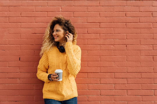 Smiling female student with long curly hair looking away while holding disposable coffee cup against red brick wall