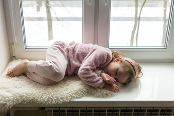 Kid lies on fur rug on windowsill at home, little girl dreams inside house in winter.