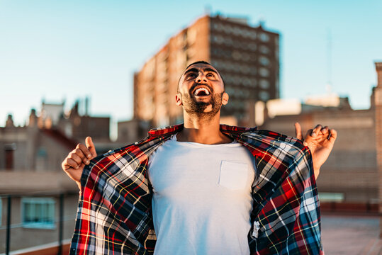 Happy man holding shirt while screaming and looking up at rooftop in city