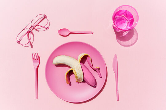 Studio shot of pink plastic tableware, glass of water, eyeglasses and pink peeled banana