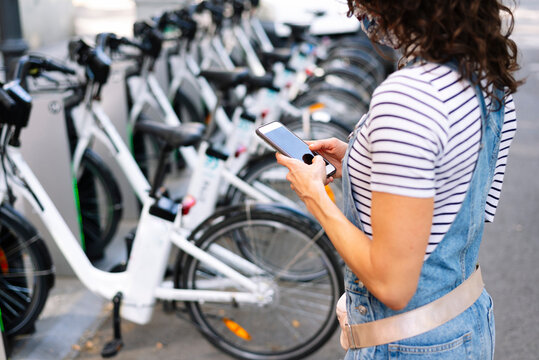 Woman standing near bicycle parking stand using smart phone in city
