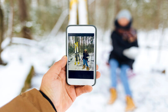 Man photographing girlfriend by skis through smart phone in forest during winter