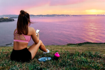 Sportswoman with smart phone sitting on grass while looking at sea against sky during sunset