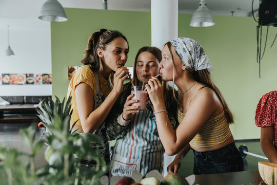 Teenage Girls Standing In Kitchen Sharing Fresh Fruit Smoothie With Drinking Straws