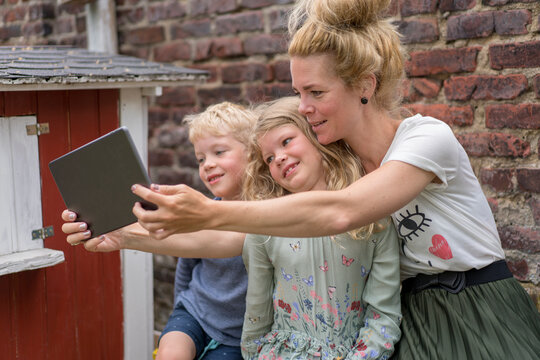 Woman taking selfie with kids through digital tablet against brick wall at back yard