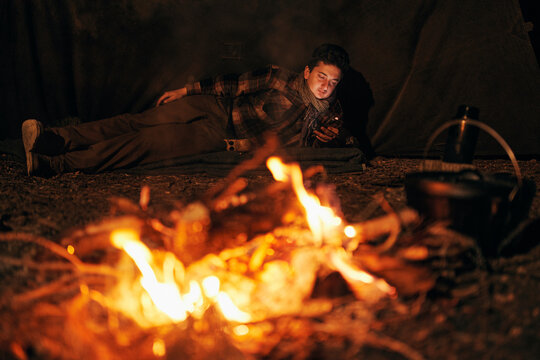 Relaxing bushcrafter using phone while lying down in forest by campfire