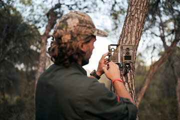 Mature male photographer positioning trail camera on tree trunk in forest