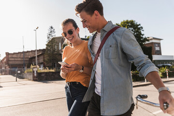 Smiling young couple looking at mobile phone while walking on street in city