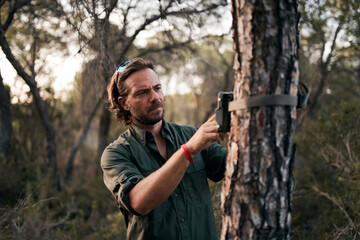 Man adjusting the settings in trail camera mounting on tree trunk in forest