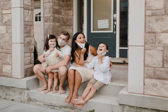 Smiling family with kids wearing protective face mask while sitting on steps outside house