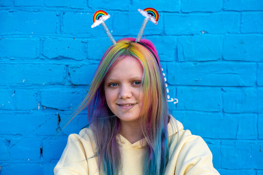 Young woman with dyed hair and rainbow alice band in front of blue wall