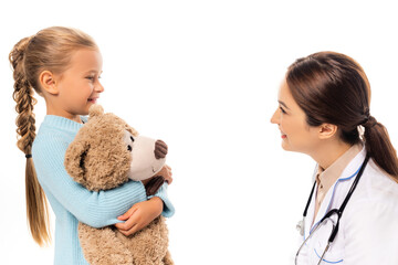 Smiling pediatrist looking at girl with soft toy isolated on white