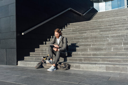Businessman using digital tablet while sitting with electric push scooter on steps in financial district