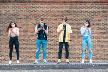 Friends maintaining safe distance while using mobile phones against brick wall in city