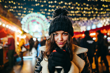 Close-up evening portrait of a cute girl at the Christmas market, standing with a cup of warming...