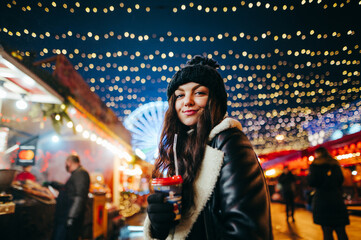 Pretty smiling girl stands on a Christmas decorated street in evening with a hot drink in her hand and poses for camera with a smile on face. Girl at the evening fair with lanterns on background.