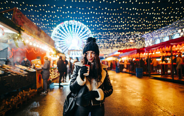 Portrait of a lovely lady in warm clothes stands at a Christmas market with beautiful light decorations, drinks a hot drink and looks away. Evening walk around the Christmas center of the city.