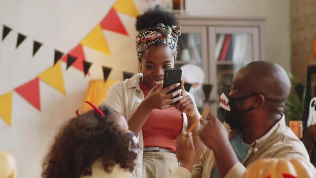 Smiling African American woman taking pictures with smartphone of little daughter and husband as they drawing blood on vampire teeth masks with felt tip pens while making Halloween costumes at home