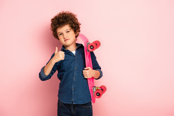 curly boy holding penny board and showing thumb up on pink