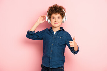 joyful and curly boy listening music in wireless headphones while showing thumb up on pink