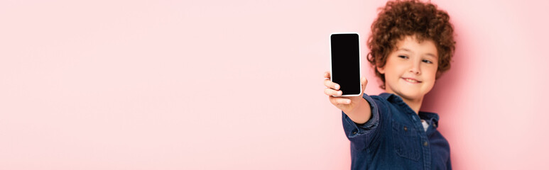 Fototapeta premium selective focus of curly boy in denim shirt holding smartphone with blank screen on pink, panoramic shot
