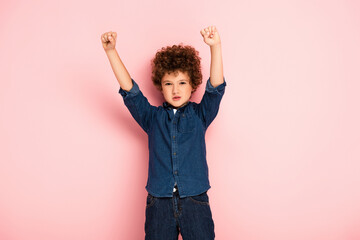 excited curly boy with hands above head and clenched fists showing winner gesture on pink