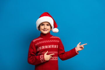 joyful boy in santa hat and red sweater pointing with fingers on blue