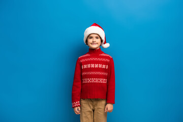 happy boy in santa hat and red knitted sweater looking at camera on blue