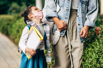 Outdoor image of a kid wearing a protective face mask going to the school looking to her mother during coronavirus. Smiling little girl with backpack and folder goes to the school holding mom's hand.