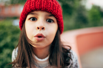 Closeup portrait of a cute little girl in red knitted hat blowing directly to the camera during resting outside after the school. Pretty child have fun outdoor.